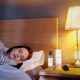 smartsleep® Pillow Spray mit dem Duft Glow, Duftspray mit Sandelholz, Labdanum, Bernstein und Patchouli zum Aufsprühen auf das Kopfkissen für Gelassenheit, Entspannung und ruhigen Schlaf auf dem Nachttisch neben einer schlafenden Frau